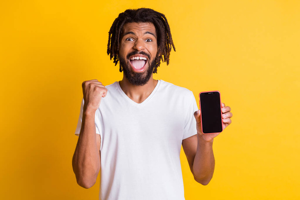 Photo of black guy hold telephone demonstrate touchscreen empty space raise fist open mouth wear white t-shirt isolated yellow color background - Φωτογραφία, εικόνα