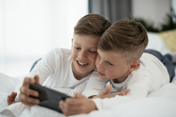 Δύο μικρά αδέρφια παίζουν παιχνίδια στο τηλέφωνο. Όμορφα αγόρια απολαμβάνουν στο σπίτι. - Φωτογραφία, εικόνα