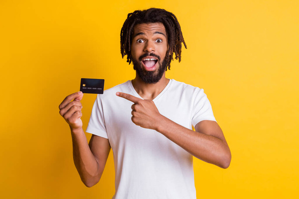 Photo of black stylish guy dreadlocks direct forefinger hold credit card open mouth wear white t-shirt isolated yellow color background - Foto, Imagem