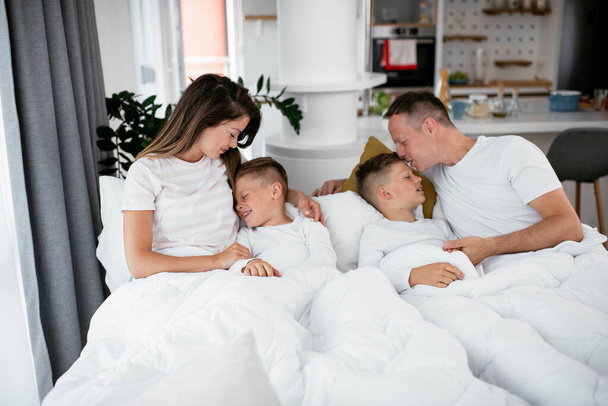 Νεαρή οικογένεια απολαμβάνει το κρεβάτι. Ευτυχισμένοι γονείς με γιους να χαλαρώνουν στο κρεβάτι. - Φωτογραφία, εικόνα