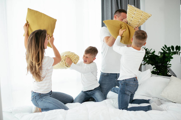 Молодые родители с сыновьями дерутся подушками на кровати. Счастливые мама и папа с мальчиками, веселящимися дома. - Фото, изображение