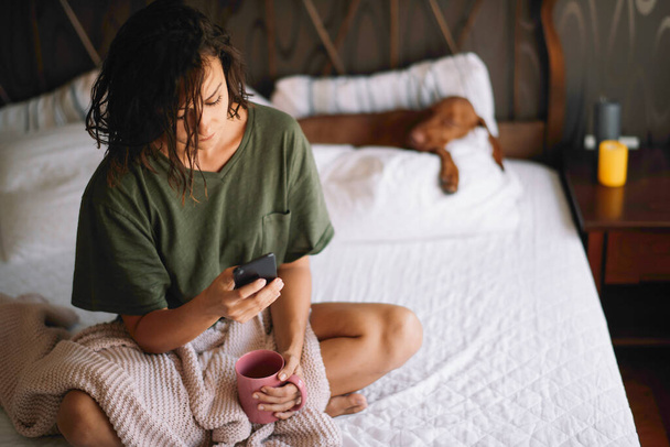 Young beautiful woman drinking coffee and using phone in bed - Fotografie, Obrázek