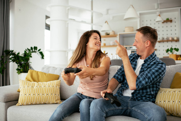 Boyfriend and girlfriend playing video game with joysticks in living room. Loving couple are playing video games at home. - Фото, изображение