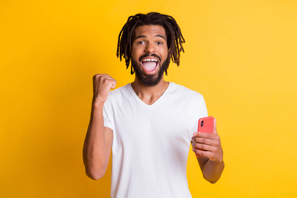 Photo of funny dark skin guy hold telephone celebrate win wear t-shirt isolated yellow color background - Foto, Bild