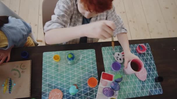 De arriba hacia abajo seguimiento de tiro de niño pequeño decorando huevo de Pascua mientras que su hermana dibujo de la imagen en el álbum en la mesa con suministros de artesanía en ella - Metraje, vídeo