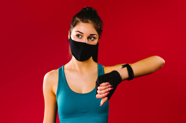 Athletic girl with a protective black mask with a fitness watch on a red background looks to the side. Sport pandemic concept - Zdjęcie, obraz