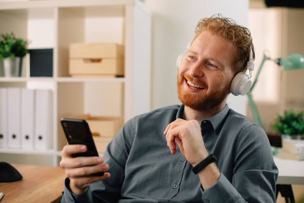Επιχειρηματίας στο γραφείο. Όμορφος άνθρωπος τζίντζερ ακούγοντας μουσική στα ακουστικά - Φωτογραφία, εικόνα