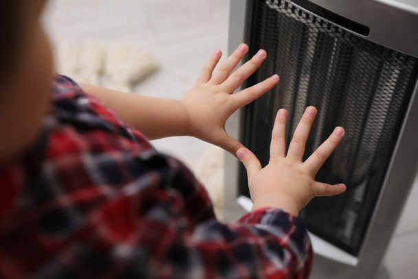 Μικρό παιδί ζεσταίνει τα χέρια κοντά σε ηλεκτρικό θερμοσίφωνα στο σπίτι, κοντινό πλάνο - Φωτογραφία, εικόνα