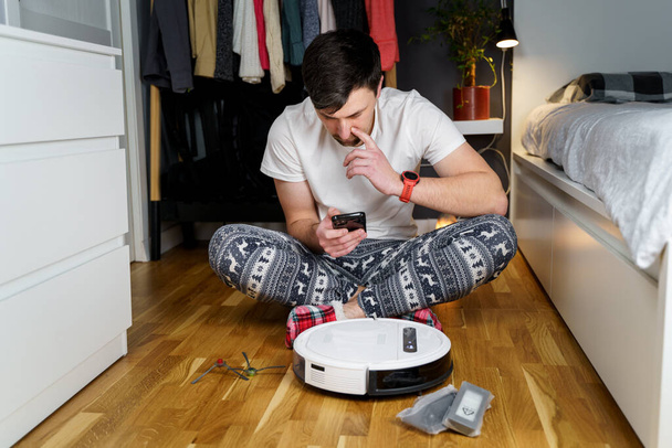 A young father in sleepwear with a winter ornament and Christmas funny socks serves a roobot vacuum cleaner for cleaning the house for the New Year and Christmas holidays. New tech for households. - Photo, Image