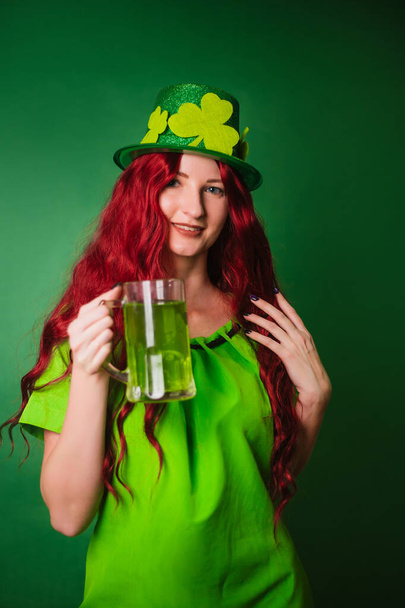 Рыжая длинноволосая девушка на зеленом фоне в шляпе с шампунем держит в руках стакан пива. Празднует День Святого Патрика. фото в студии - Фото, изображение