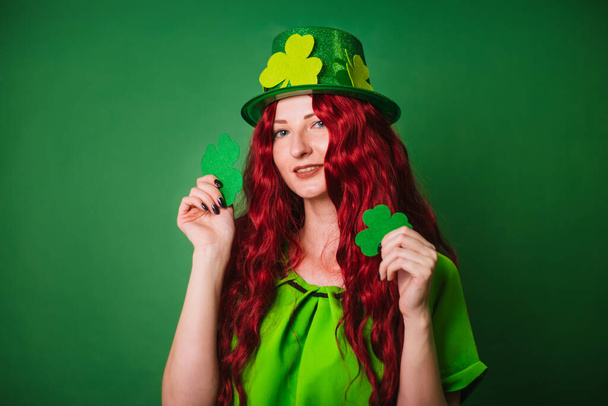 緑の背景をした赤髪の長髪の少女は、手に三枚葉の木を持っている。聖パトリックの日を祝う。スタジオでの写真 - 写真・画像