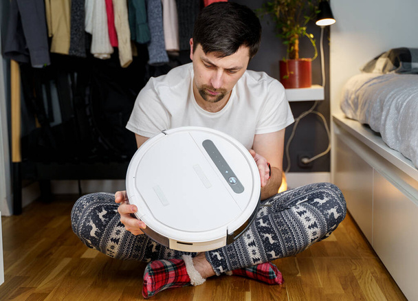 Caucasian young man in christmas pajamas doing vacuum cleaner robot maintenance. Male in winter patterned sleepwear and red socks slippers starts automatic vacuum cleaner. Robot vacuum cleaner repair. - Photo, Image