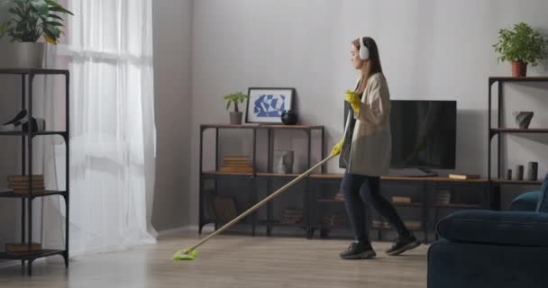 Hauswirtschaft und Aufräumen der Wohnung, junge fröhliche Frau wäscht Boden mit Wischmopp und tanzt, hört Musik über Kopfhörer - Filmmaterial, Video