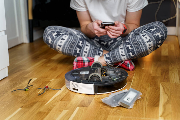 Young caucasian man doing cleaning for the holiday christmas in winter. A male in funny home clothes, pajamas with winter ornament collects and sets up a robot vacuum cleaner at home on the floor. - Valokuva, kuva