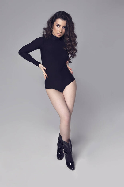 Full length front portrait of a beautiful girl with brunette curly hair and makeup, in a black bodysuit posing in the studio, grey background. Vertical view. - Photo, Image