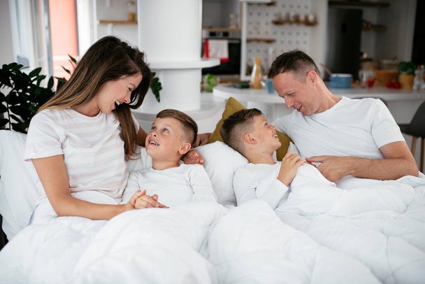 Молода сім'я насолоджується в ліжку. Щасливі батьки з синами відпочивають у ліжку
. - Фото, зображення