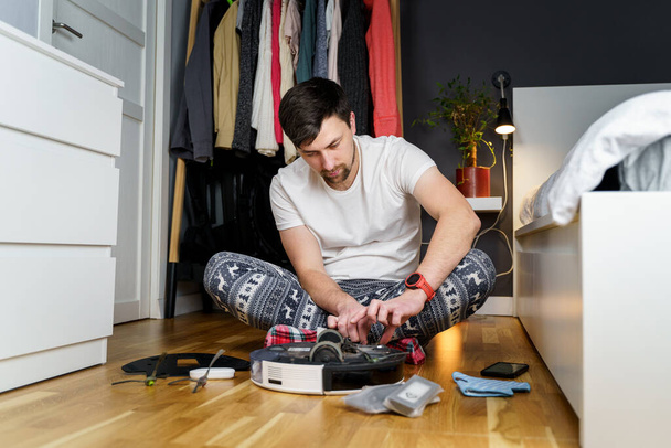 Fiatal kaukázusi férfi takarít az ünnepekre télen. Egy férfi vicces otthoni ruhákban, pizsama téli dísszel összegyűjti és felállítja a robot porszívót otthon a padlón. - Fotó, kép