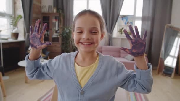 Portret van een schattig klein meisje met handen met verf erop, kijkend naar de camera en vrolijk lachend terwijl ze thuis poseert - Video
