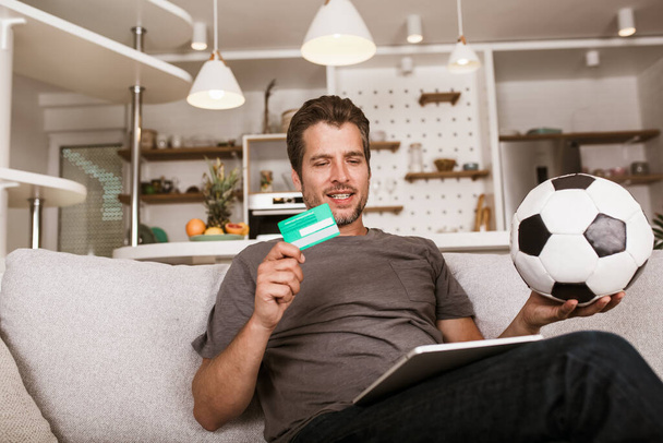 Junger Fußballfan, der auf der Wohnzimmercouch ein Fußballspiel im Fernsehen verfolgt. Er hält eine Kreditkarte und ein digitales Tablet für Online-Wetten. - Foto, Bild