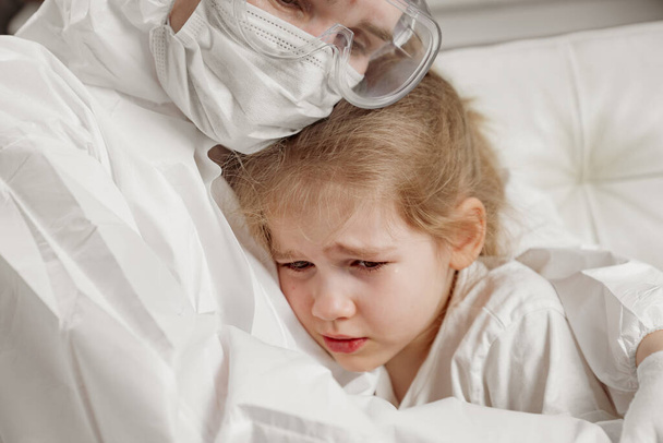 dokter in beschermend pak, masker, handschoenen en bril kalmeert klein ziek meisje. - Foto, afbeelding
