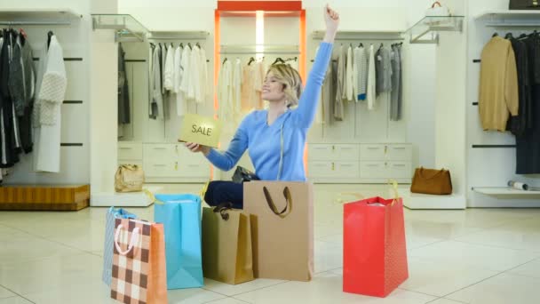 Black Friday and shopping sale. Happy young woman holding sale text sign and dancing surarronded with colorful bags full of purchases . Attractive woman smiling at camera with paper bags around. 4k - Footage, Video