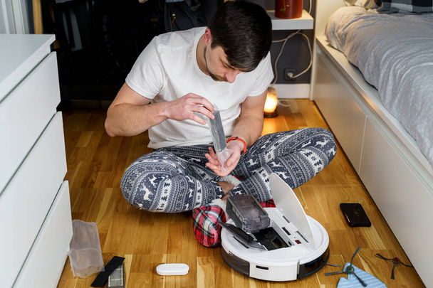 クリスマスのパジャマ姿の白人青年が掃除ロボットのメンテナンスをしている。冬のパターン化された眠気や赤い靴下スリッパの男性は自動掃除機を開始します。ロボット掃除機修理 - 写真・画像