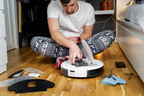 Hombre joven caucásico en pijama de navidad haciendo mantenimiento de robot aspiradora. Hombre en invierno modelado ropa de dormir y calcetines rojos zapatillas comienza aspiradora automática. Robot aspiradora reparación - Foto, imagen