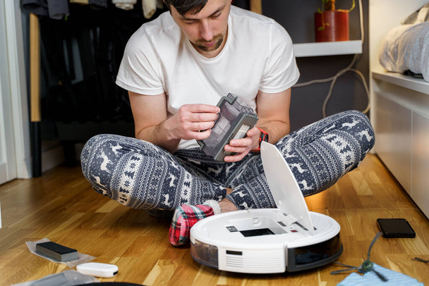 Caucasian young man in christmas pajamas doing vacuum cleaner robot maintenance. Male in winter patterned sleepwear and red socks slippers starts automatic vacuum cleaner. Robot vacuum cleaner repair - Photo, Image