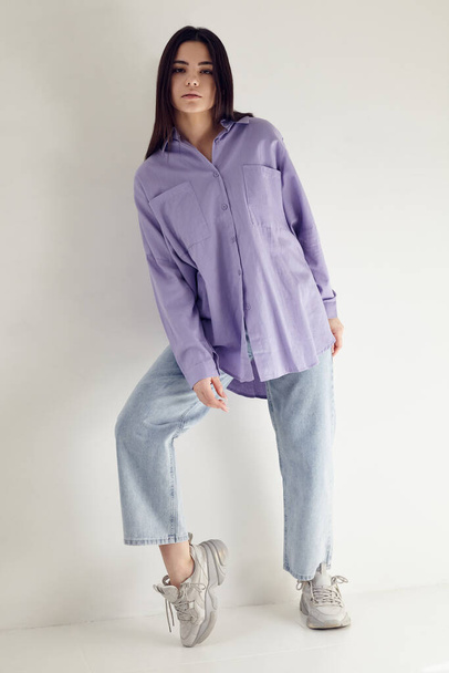 beautiful brunette in a lilac shirt and blue jeans ina light background - Photo, image