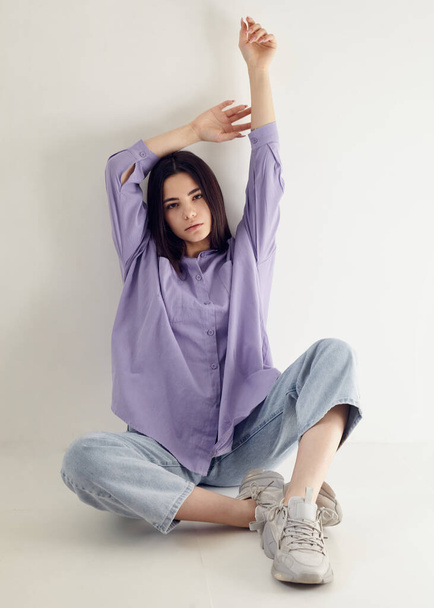 beautiful brunette in a lilac shirt and blue jeans ina light background - Photo, Image