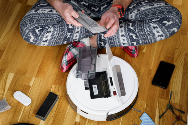 クリスマスのパジャマ姿の白人青年が掃除ロボットのメンテナンスをしている。冬のパターン化された眠気や赤い靴下スリッパの男性は自動掃除機を開始します。ロボット掃除機修理 - 写真・画像