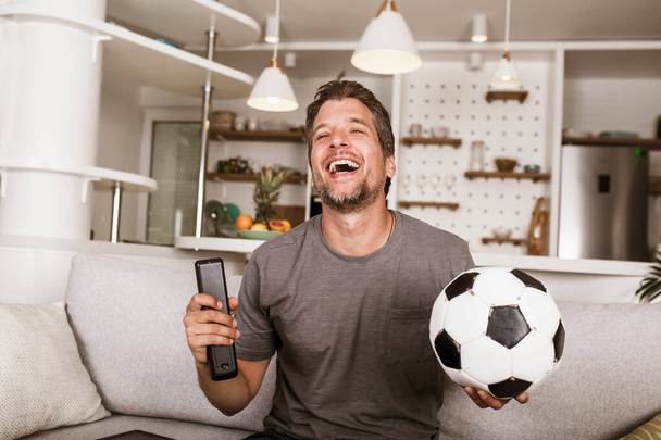 Młody fan piłki nożnej człowiek ogląda mecz piłki nożnej w telewizji na kanapie w salonie. - Zdjęcie, obraz
