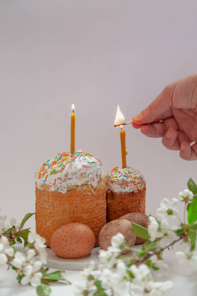 Two Easter cakes and eggs stand on a white background. Candles are inserted into the Easter cakes. A hand lights a candle with a match. In the foreground is a tree branch with white flower - Фото, изображение