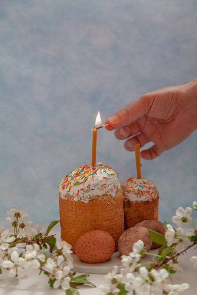 Two Easter cakes and eggs stand on a blue background. Candles are inserted into the Easter cakes. A hand lights a candle with a match. In the foreground is a tree branch with white flower - Фото, изображение