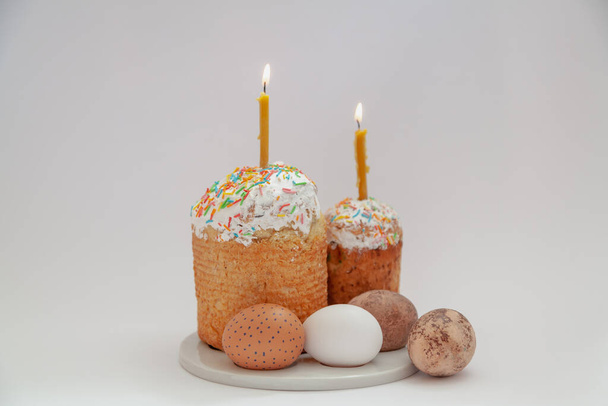 Easter still life. Two Easter cakes and eggs stand on a round stand on a white background. Burning candles are inserted into Easter cake - Фото, изображение