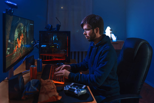 Joueur professionnel concentré jouant le jeu, prenant part au tournoi en ligne assis à l'ordinateur à la maison. Faible luminosité, couleur néon, vue de côté. Joueur professionnel cybersport entraînement jeu en ligne sur son PC - Photo, image