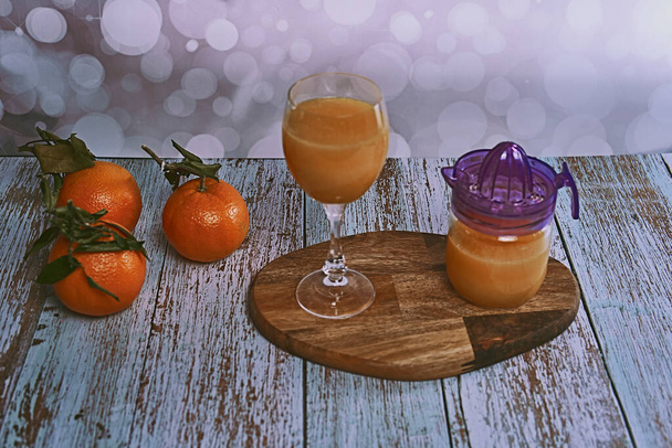 Verre de jus d'orange avec presse-agrumes en verre sur table en bois, diverses oranges sur sol en bois et fond lumineux - Photo, image