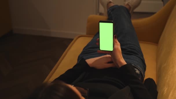 Back view. Woman using phone green mock-up screen chromakey lying on sofa couch at home office. Girl using smartphone, Browsing Internet, Watching Content. Woman has video call chatting on phone. - Footage, Video