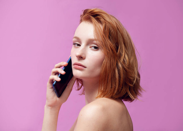 beautiful redhead girl with a phone in her hands on a pink background - Foto, imagen