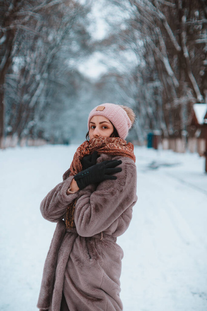 Portrait of a young stylish caucasian woman posing in park hiding behind a scarf in winter in a long eco-friendly fur coat under a snowfall. Concept of winter season. People enjoy real winter - Photo, Image