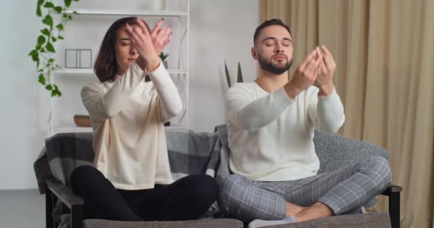 Šťastný pár dělá ranní trénink doma moderní obývací pokoj na gauči. Kavkazská žena a muž pohybující se ruce dělat stejný pohyb s rukama sedí v lotosu pózovat medituje, duchovní praktiky lekce - Záběry, video