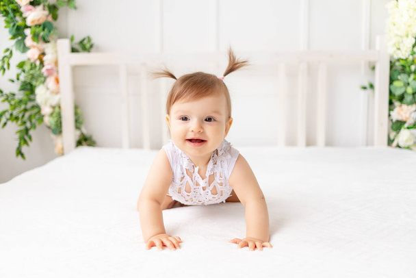 divertida niña de seis meses de edad sentado en una hermosa habitación brillante en una cama blanca en un traje de encaje mira hacia otro lado y sonríe - Foto, imagen