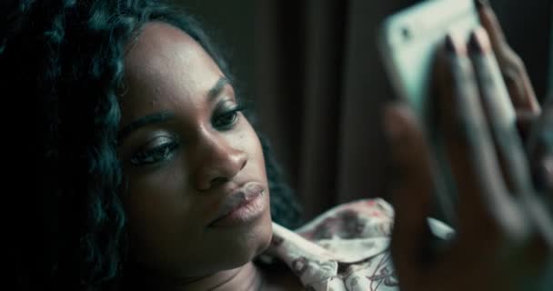 Gyönyörű afro-amerikai nő, göndör afro frizurával a kanapén. Fiatal nő néz valamit a telefonján, és jó szórakozást. Hihetetlen, egzotikus külsejű lány. 4K videó - Felvétel, videó
