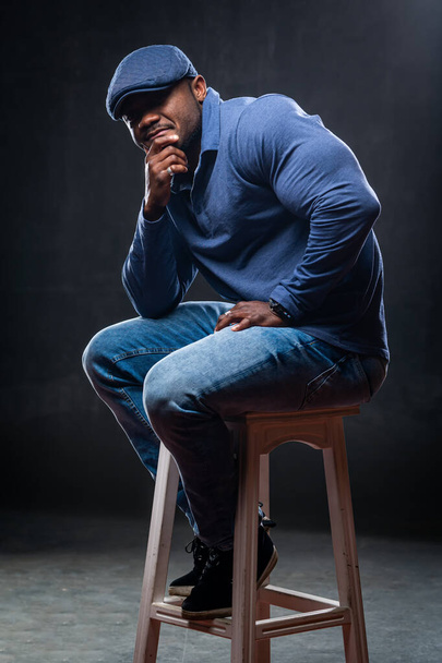 Thoughtful big muscular man sits on high bar chair. Male wears casual blue clothes. Studio photo. - Foto, Imagem