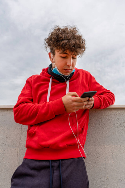 Mladý atraktivní muž s kudrnatými vlasy, oblečený v červené mikině, používající mobilní telefon na terase domu s maskou, aby se chránil před koronavirem. Nový normální koncept - Fotografie, Obrázek