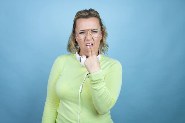 Молода біла жінка в навушниках на шиї на синьому фоні огидною рукою всередині рота
 - Фото, зображення