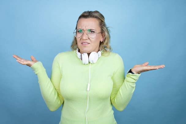 Mujer caucásica joven con auriculares en el cuello sobre fondo azul expresión despistada y confusa con los brazos y las manos levantadas - Foto, imagen