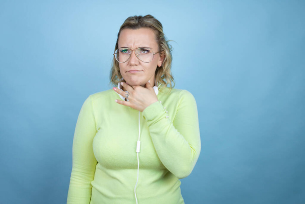Mulher branca jovem usando fones de ouvido no pescoço sobre fundo azul tocando pescoço doloroso, dor de garganta para gripe, cravo e infecção - Foto, Imagem