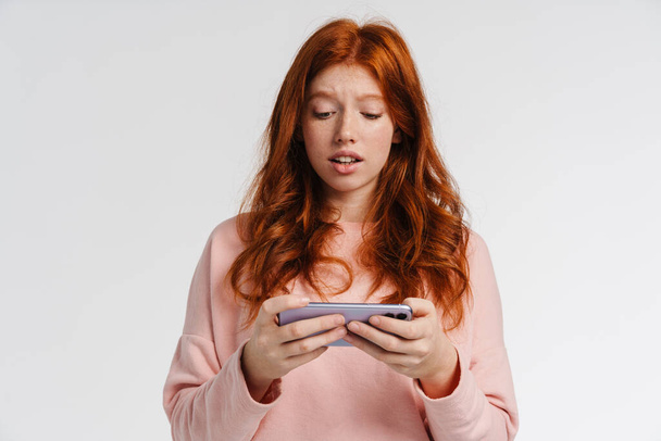 Ginger beautiful focused girl playing online game on cellphone isolated over white background - Fotografie, Obrázek