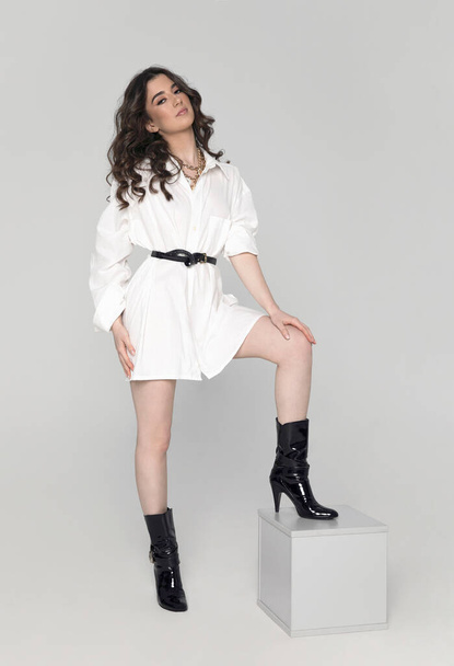 Full length image of a young brunette curly woman with hairstyle and makeup, in long white shirt and high heel posing in studio. Vertical view. - Photo, Image